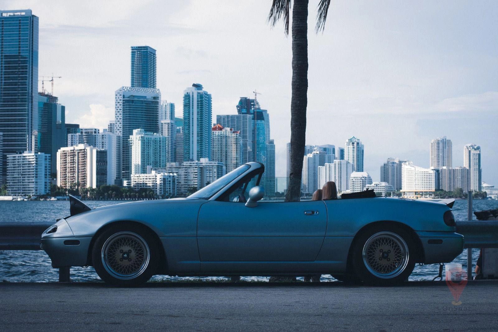A blue Miata with its top down in front of Miami’s waterfront skyline